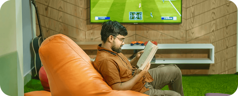 Person relaxing in an orange beanbag reading while watching a soccer match on TV