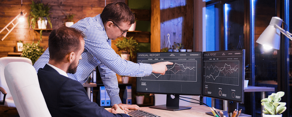 Colleagues reviewing annual report dashboards on dual monitors at night