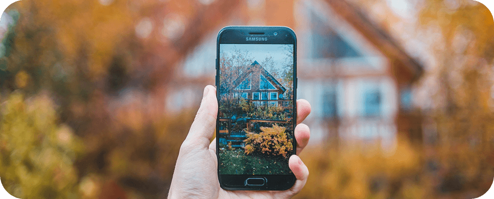 Hand holding a smartphone displaying a modern house against an autumn landscape