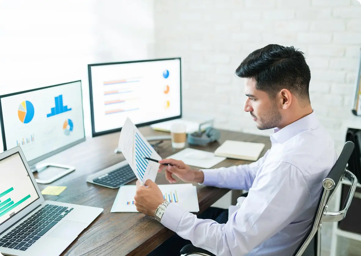 Professional comparing printed reports with data charts on dual monitors