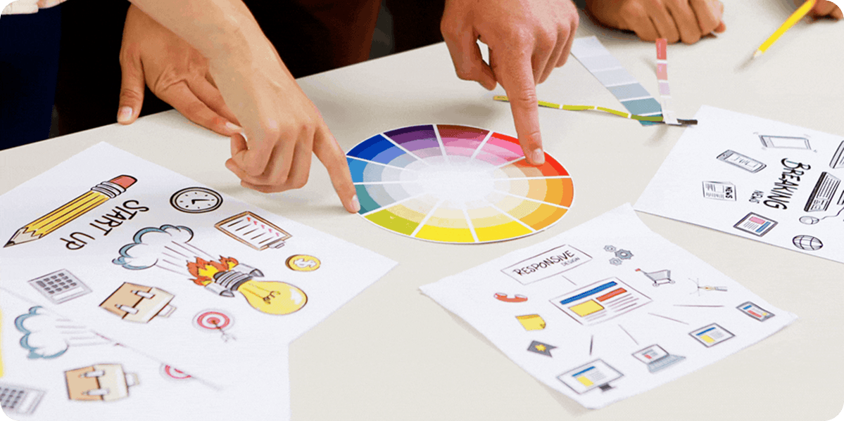 Hands collaborating around a color wheel with design sketches on a table