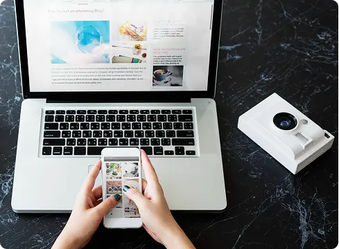 Hands browsing a website on a phone next to a laptop and camera