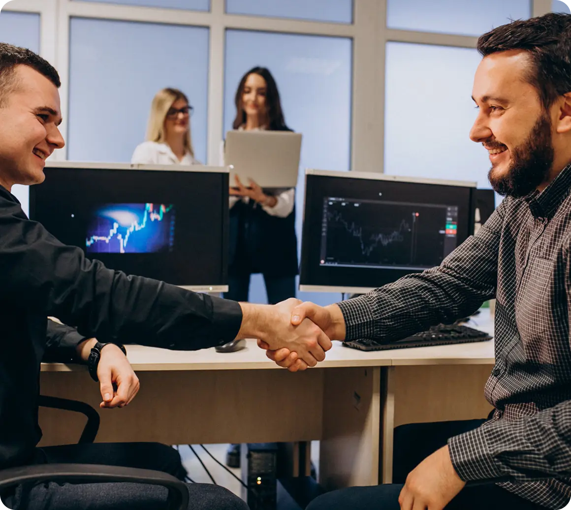 Two professionals shaking hands in an office with market data on screens